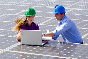 two engineers wearing hard hats looking at a laptop while standing among solar panels